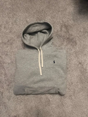 Grå hoodie från Ralph Lauren - Snygg grå hoodie från Ralph Lauren med klassisk huva och vita dragsnören. Tröjan har en broderad marinblå logga på bröstet och Tillverkad i mjukt bomullsmaterial och har ribbade muddar vid ärmslut och nederkant.