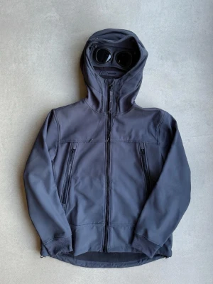CP company softshell jacka - Snygg mörkgrå vindjackan - C.P. Company med ikoniska inbyggda glasögon i huvan. Jackan har dragkedja framtill, två vertikala fickor med dragkedja och muddar vid ärmsluten. softshell jacka.