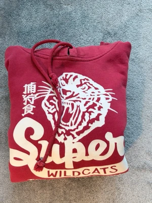 Superdry Hoodie - Snygg röd hoodie från Super Tokyo Wildcats med stor vit tigerprint och text på bröstet. Klassisk känguruficka, ribbade muddar och dragsko i huvan. Skön och casual stil, perfekt till jeans eller joggers. Materialet känns mjukt och tjockt, troligen bomullsblandning.