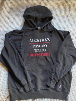 vintage Svart hoodie med Alcatraz-tryck y2k - Svart hoodie med huva och dragsnören. På bröstet står det 'ALCATRAZ PSYCHO WARD OUTPATIENT' i vitt och rött tryck. Klassisk känguruficka framtill och ribbade muddar. Perfekt för dig som gillar statement-plagg och streetwear.