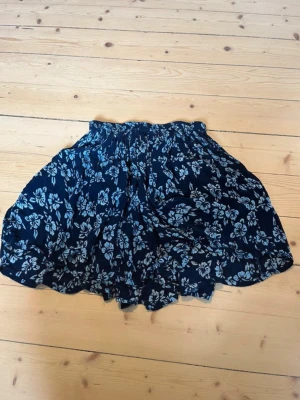 Blommig blå kortkjol från Indiska - Supersöt kortkjol från Indiska i mörkblått med ljusblått blommönster. Kjolen har en luftig och flowy passform med resår i midjan, perfekt för varma dagar. Materialet känns lätt och bekvämt mot huden. OBS! Storleksmärkt 146/152 men passade mig som XS. 