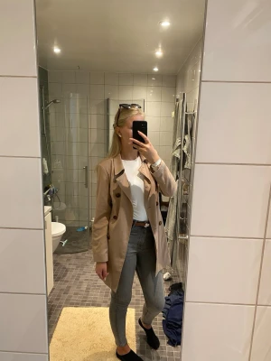 Beige trenchcoat med knytskärp - Snygg beige trenchcoat med klassisk dubbelknäppning och knytskärp i midjan. Jackan har stora bruna knappar och en stilren krage. Perfekt för lager-på-lager och passar till jeans eller dressade byxor. Materialet ser ut att vara bomull eller polyester.