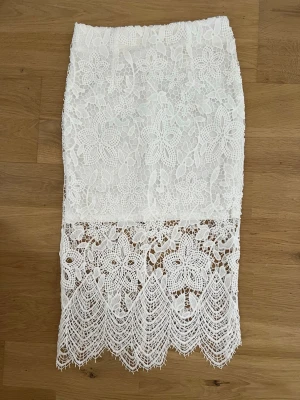 Vit spetskjol med blommigt mönster - Supersnygg vit långkjol i spets med blommigt mönster och vacker volangkant nedtill. Kjolen har en feminin och elegant look med genombrutna detaljer och hög midja. Perfekt för dig som vill sticka ut med en romantisk stil.