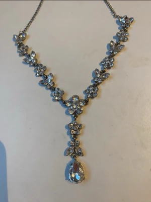 Silvrigt halsband med gnistrande stenar - Säljer ett elegant halsband i silverfärg med gnistrande sopade stenar i blommönster och en stor droppformad sten som hänge. Kedjan är tunn och smycket har en lyxig känsla med detaljerade blomdekorationer. Perfekt för dig som vill sticka ut med något glittrigt.