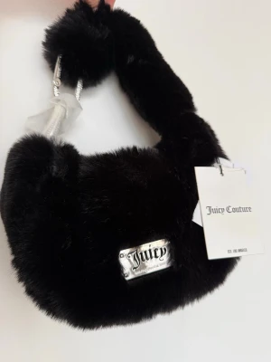 Svart fluffig handväska Juicy Couture - Supersnygg svart handväska från Juicy Couture i mjukt, fluffigt fuskpälsmaterial. Väskan har en silvrig metallogo framtill och kort axelrem med kedjedetaljer. Perfekt accessoar för att lyfta din outfit.