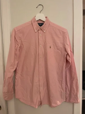 Rosa skjorta från Polo Ralph Lauren - Säljer en klassisk rosa skjorta från Polo Ralph Lauren med button-down krage och broderad logga på bröstet. Skjortan har långa ärmar och knappar framtill. Perfekt för dig som gillar stilrena plagg med ikonisk design.