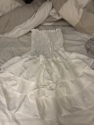 Vit axelbandslös kortklänning - Söt vit axelbandslös kortklänning med smockad överdel och volangig kjol. Klänningen har en luftig känsla och passar perfekt till varma dagar. Materialet känns lätt och mjukt, troligen bomull eller liknande. Enkel och stilren design.