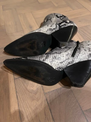 Twist & Tango  boots i ormskinnsmönster - Snygga cowboy boots med spetsig tå och coolt ormskinnsmönster i svart, grått och vitt. Bootsens ovandel har dragkedja på sidan och klacken är kraftig och vinklad. Perfekta för dig som vill sticka ut med din stil. Äkta skinn, storlek 39. Perfekt skick.