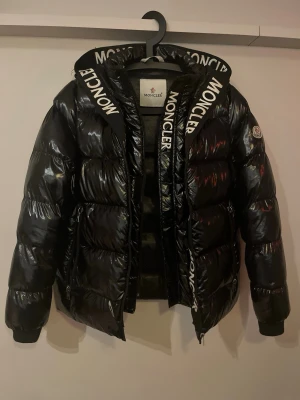 Svart Moncler dunjacka med logoband - Snygg svart dunjacka från Moncler med glansig finish och stora, breda logoband med Moncler-tryck. Jackan har hög krage, dragkedja och klassisk puffer-design som håller dig varm under kalla dagar. Perfekt för dig som vill sticka ut med en exklusiv vinterjacka.