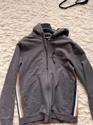 Grå zip hoodie från BOSS - Snygg grå hoodie från BOSS med dragkedja framtill och huva med dragsko. Diskret BOSS-logga på bröstet och coola ränder i beige, svart och vitt längs sidorna. 