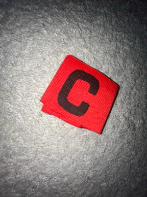 Rött kaptensbindel med svart C - Säljer en röd kaptensbindel med ett stort svart C på framsidan. Perfekt för dig som vill ta ledarrollen på planen. Bindeln är elastisk och passar de flesta armstorlekar. Enkel att ta på och av.