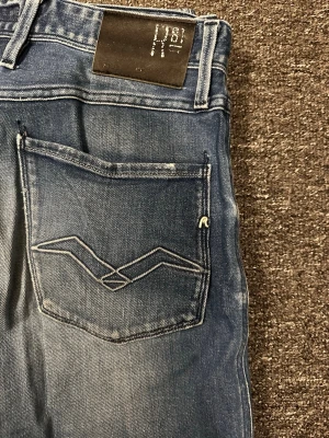 Replay anbass - Snygga blå slim fit jeans från Replay med slitna detaljer och ljusa tvättade partier framtill. Klassisk femficksmodell med broderad bakficka och Replay-logga på linningen. Perfekta för en avslappnad och trendig stil.