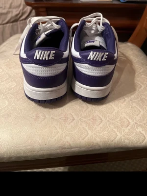 Nike Dunk Low vit/lila sneakers - Snygga Nike Dunk Low sneakers i vitt och lila med klassisk design. Skorna har vita snören, perforerad tåbox och tydlig Nike-logga bak. Ovandelen är i lä soppa och textil, vilket ger både stil och komfort. Perfekta för dig som vill ha en clean och sportig look.