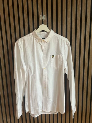 Vit skjorta från Lyle & Scott - Klassisk vit skjorta från Lyle & Scott med button-down krage och bröstficka med den ikoniska drawloggan i gult och svart. Skjortan har långa ärmar och knappar framtill. Perfekt till en clean och stilren look.