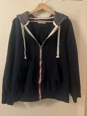 Pier One Mörkblå hoodie med dragkedja - Snygg Pier One mörkblå hoodie med dragkedja och två fickor framtill. Grå huva med vita snören och rödvit detalj längs dragkedjan. Perfekt för en avslappnad stil och enkel att matcha med jeans eller joggers.