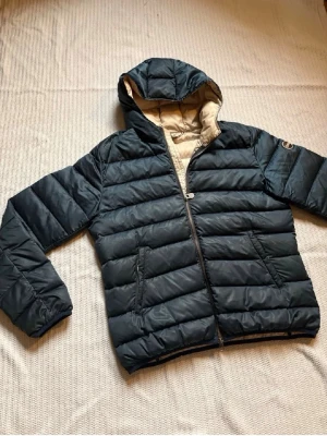 Mörkblå colmar jacka - Snygg mörkblå pufferjacka med beige innerfoder och huva. Jackan har dragkedja framtill, två sidofickor och ett märke på ärmen som visar loggan. Perfekt för vinterdagar men även nu för lite kyligare vårväder. Köpt på plick. Små hål i armarna finns men syns inte mycket, därav det lägre priset. Jag upplever jackan som lite stor men det är min uppfattning, det är inte så synligt dock. 