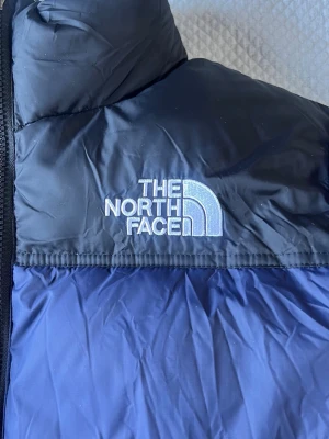 Blå dunjacka från The North Face - Snygg blå dunjacka från The North Face med broderad logga på bröstet. Jackan har en tvåfärgad design i mörkblått och svart, dragkedja framtill och är perfekt för kalla vinterdagar. Tillverkad i slitstarkt material och fylld med dun för extra värme.