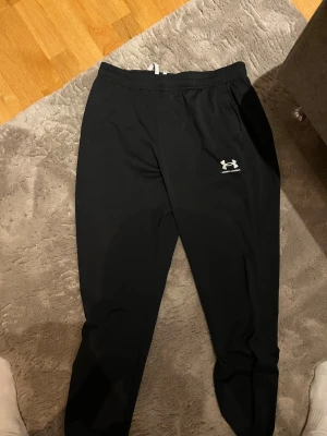 Svarta mjukisbyxor från Under Armour - Säljer ett par svarta mjukisbyxor från Under Armour med dragsko i midjan och logga på vänster lår. Byxorna har en slim fit passform och är tillverkade i ett mjukt och stretchigt material som är perfekt för chill eller träning.