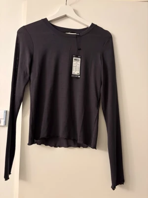 Grå långärmad topp med vågig kant prislapp  - En grå långärmad topp med rund hals och vågiga kanter vid ärmslut och nederkant. Tunn och mjuk kvalitet som passar perfekt till jeans eller kjol. Enkel och stilren design som är lätt att matcha. Prislapp kvar strl M 