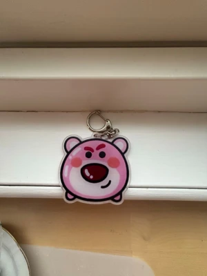Nyckelring med rosa björnfigur - Söt nyckelring med en tecknad rosa björn med runda former och röda detaljer. Nyckelringen har en metallhake som gör den enkel att fästa på väskan eller nycklarna. Tillverkad i plast med tryckt motiv, perfekt för att ge din stil en lekfull touch.