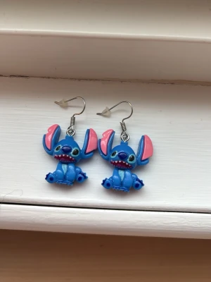 Blå Stitch örhängen med krokar - Säljer ett par unika örhängen med motiv av Stitch från Disney. Örhängena är blå med rosa detaljer på öronen och har krokar i metall. Perfekta för dig som älskar tecknade figurer och vill ha något lekfullt till din stil.