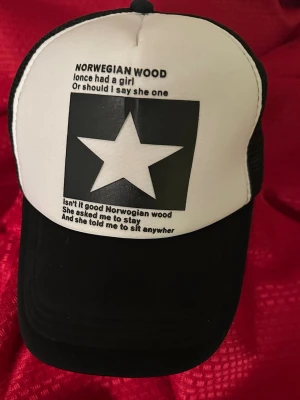 Svartvit truckerkeps med stjärntryck - Säljer en svartvit truckerkeps med stort stjärntryck och texten 'Norwegian Wood' på framsidan. Kepsen har svart nät på sidorna och justerbar baksida. Perfekt för dig som gillar unik och grafisk stil.