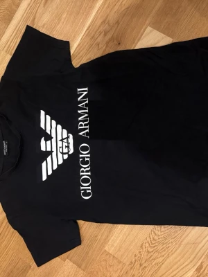 Svart t-shirt från Giorgio Armani - Snygg svart t-shirt från Giorgio Armani med stort vitt logotryck och text på framsidan. Klassisk passform med rund hals och korta ärmar. Perfekt för dig som gillar stilrena märkeskläder.