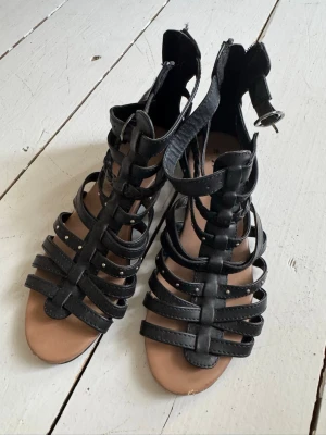 Svarta gladiatorsandaler med spänne - Säljer ett par svarta gladiatorsandaler med flera remmar och öppen tå. Sandalerna har dragkedja bak och ett justerbart spänne vid ankeln. Tillverkade i konstläder med platt sula, perfekta för en trendig och avslappnad look.