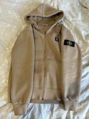 Beige hoodie med dragkedja Stone Island - Snygg beige hoodie från Stone Island med dragkedja framtill och klassiska logopatchar på bröstet och ärmen. Hoodien har huva med dragsko sopm gör den extra chill och muddar vid ärmslut och nederkant. Perfekt för en avslappnad streetwear-look.