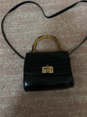 Svart croco handväska med bambuhandtag - Stilren svart handväska i croco-mönstrat konstläder med unikt bambuhandtag och guldfärgat spänne. Väskan har både axelrem och handtag, samt ett rymligt fack inuti. Perfekt för dig som gillar trendiga detaljer och klassisk look.