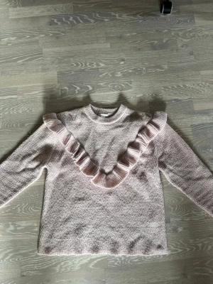 Ljusrosa stickad tröja med volang - Supersöt ljusrosa stickad tröja från Gina Tricot med volangdetalj i V-form framtill. Tröjan har lång ärm, rund halsringning och ett diskret vågmönster i stickningen. Perfekt för dig som gillar gulliga detaljer och mjuka material.