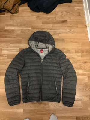 Grå dunjacka med huva från Colmar - Snygg grå dunjacka från Colmar med huva och dragkedja framtill. Jackan har två dragkedjeförsedda fickor och Colmar-logga på ärmen. Perfekt för våren och har en stilren, quiltad design. Defekt finns på högra armbågen där det är ett litet hål som kanske är 1 cm men det går att fixa med rätt grejer.