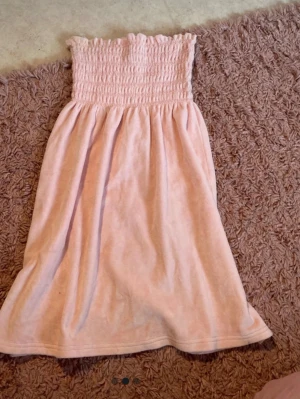 Ljusrosa klänning Juicy Couture - Supersöt ljusrosa kortklänning från Juicy Couture i mjuk frotté. Klänningen är axelbandslös med smock upptill och har en avslappnad passform. Perfekt för sommarens varma dagar eller som strandklänning.