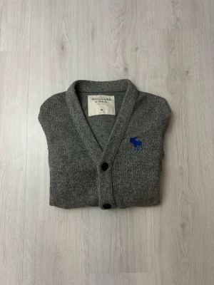 Grå kofta från Abercrombie & Fitch - Mysig grå kofta från Abercrombie & Fitch med blå logga broderad på bröstet. Koftan har v-ringning, knäppning med svarta knappar och två fickor framtill. Ribbstickade muddar vid ärmslut och nederkant. Perfekt för lager på lager och chill stil.