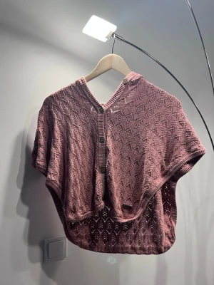 Rosa stickad poncho med huva - Säljer en ljusrosa stickad poncho med korta ärmar och huva. Ponchon har dekorativa knappar framtill och ett fint hålmönster över hela plagget. Perfekt för lager på lager och ger en avslappnad men snygg look.