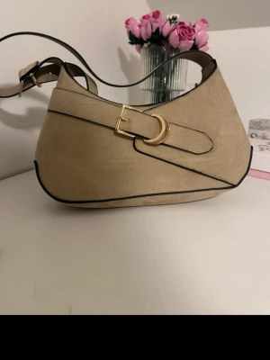 Beige axelremsväska med gulddetalj - Snygg beige handväska i mockaimitation med svart kantband och guldfärgat D-spänne. Väskan har en justerbar axelrem och unik form som ger en trendig look. Perfekt för dig som vill ha en stilren accessoar med det lilla extra.