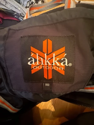 Svart dunjacka från áhkká Outdoor - Ord pris 600 sekSnygg svart dunjacka från áhkká Outdoor med quiltad design och dragkedja framtill. Jackan har en diskret logga på bröstet och är perfekt för kalla vinterdagar. Tillverkad i slitstarkt material som håller värmen bra.