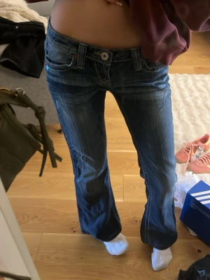 Blå bootcut jeans från True Religion - Säljer ett par blå bootcut jeans från True Religion med klassiska kontrastsömmar och snygga detaljer på bakfickorna. Jeansen har låg midja och är tillverkade i denim med lätt slitning. Perfekt för dig som gillar en avslappnad och trendig look.