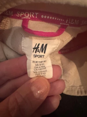Fleecejacka H&M Sport rosa/vit - Mysig fleecejacka från H&M Sport i ljusbeige med rosa ärmar. Jackan har hel dragkedja framtill, bröstficka med dragkedja och hög krage. Perfekt för träning eller som ett extra lager under kalla dagar.