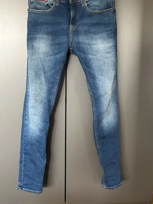 Tommy Jeans Austin Slim Tapered - Snygga blå jeans från Tommy Jeans i modellen Austin Slim Tapered. Jeansen har en smal passform med avsmalnande ben och klassisk femficksdesign. Tillverkade i slitstark denim med snygga slitningar och ljusare partier för en trendig look.