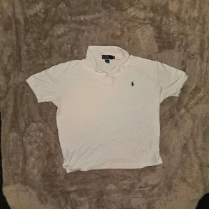Vit pikétröja från Ralph Lauren - Klassisk vit pikétröja från Ralph Lauren med korta ärmar och den ikoniska broderade loggan på bröstet. Tröjan har krage och knappar vid halsen. Perfekt till jeans eller shorts för en clean och stilren look. Har fadade fläckar vid armen som inte syns så mycket vid användning har också två små blå fläckar vid knapparna men dom är också svåra att märka av om man inte adaptivt letar efter det