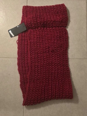 Vinröd stickad halsduk från New Yorker  - Säljer en chunky vinröd halsduk snood från New Yorker i grovstickad design. Halsduken är tillverkad i 100% polyester och har en mjuk och varm känsla. Perfekt för att addera färg till din outfit under kyliga dagar.oanvänd och i väldigt bra skick 