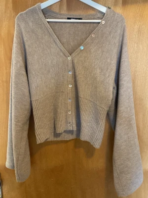 Beige kofta från Gina Tricot - Beige kofta från Gina Tricot med v-ringning och knappar framtill. Koftan har ribbstickad nederdel och långa ärmar. Perfekt för lager-på-lager och enkel att matcha med olika outfits. Använd max 2 gånger.