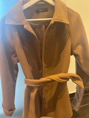 Kamelbrun kappa från Gina Tricot - Stilren kamelbrun kappa från Gina Tricot med knytskärp i midjan och klassisk krage. Kappan har knappar framtill och raka ärmar. Perfekt för dig som vill ha en tidlös och trendig look till hösten eller vintern.