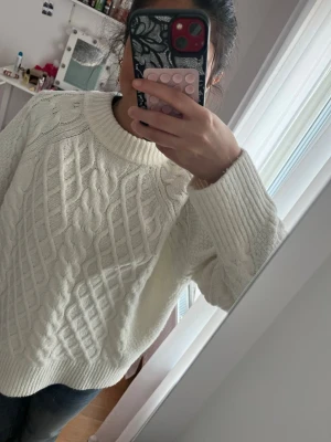 Vit kabelstickad tröja - Säljer en vit kabelstickad tröja med klassiskt mönster och rund halsringning. Tröjan har ribbade muddar vid ärmslut och nederkant samt långärmad design. Materialet ser ut att vara ull eller ullblandning, perfekt för kyliga dagar.