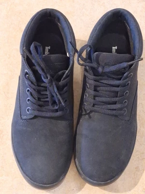 Svart Timberland sneakers - Snygga svart sneakers från Timberland med klassisk låg profil. Skorna har ovandel i mocka och textil, matchande mörkblå snören och diskret logga på innersulan. Perfekta för dig som gillar stilrena och tidlösa sneakers. 