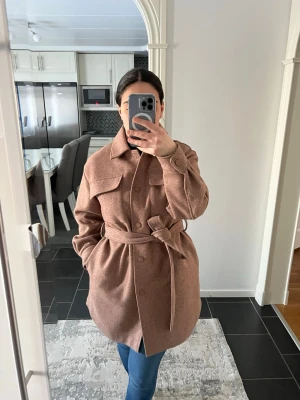 Brun kappa med knytskärp - Stilren brun kappa i ullmix med knytskärp i midjan och stora bröstfickor. Kappan har klassisk krage, knäppning framtill och en rak, något oversized passform. Perfekt för dig som gillar en trendig och avslappnad look. Nypris: 669kr. Pris går att diskuteras. 