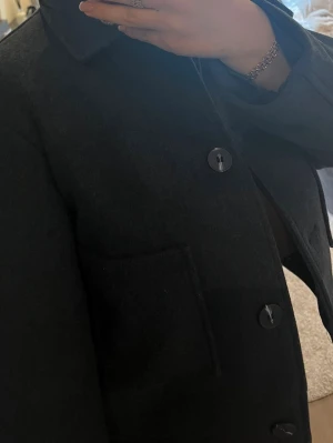 Marinblå  ullkappa med stora knappar - Marinblå svart k rendered ullkappa med4 stora knappar framtill och klassisk krage. Kappan har en rak passform och en synlig bröstficka. Perfekt för dig som vill ha en enkel men snygg look till hösten eller vintern.