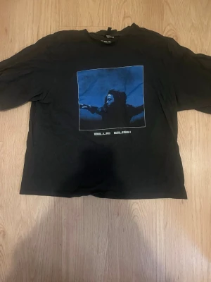 Svart Billie Eilish t-shirt med blå tryck - Cool svart t-shirt från Billie Eilish med blått tryck på framsidan och låtlista på baksidan. T-shirten har en klassisk passform och är kortärmad, perfekt för dig som gillar musik och streetstyle.