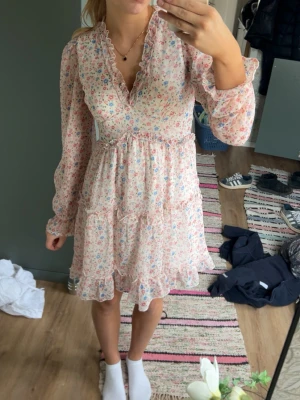 Blommig kortklänning med volang - Söt kortklänning med volangdetaljer och v-ringning. Klänningen har långärmade, lätt puffade ärmar och ett romantiskt blommönster i rosa, blått och grönt på ljus botten. Tillverkad i ett tunt, luftigt material som känns perfekt för vår och valuesommar.